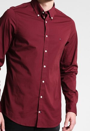 Mann trägt ein langärmeliges, knöpfbares weinrotes Hemd mit kleinem Logo auf der linken Brust, steht vor einem einfarbigen Hintergrund.