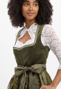 Frau in einem grünen Samtkleid mit großer Schleife und weißer Spitzenbluse mit floralem Muster, die leicht zur Seite schaut, mit entspannten Armen.