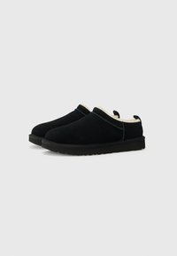 Svarte semskede slip-on sko med myk beige fôr, rund tå og teksturerte gummisåler, med en bakflik for enkel påtaking.