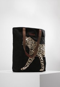 Tote bag en toile noire avec anses en cuir marron, ornée d'un motif léopard en sequins dorés sur un côté. Spacieux avec une forme structurée.