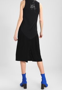 Czarny bezrękawnik z zaokrąglonym dołem i graficznym wzorem na plecach, w połączeniu z czarną spódnicą do kolan i niebieskimi botkami za kostkę.