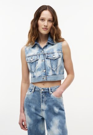 Ung kvinde iført ærmeløs cropped acid-wash denimjakke med knaplukninger og matchende højtaljede acid-wash jeans, hånd i lommen.