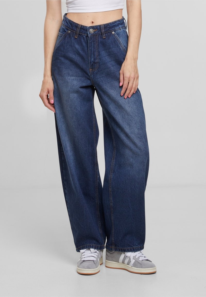 Personne portant un jean denim bleu foncé à jambes larges, un top court blanc et des baskets grises avec des lacets blancs, debout sur un fond gris uni.
