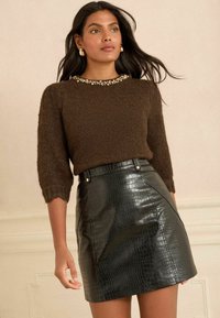 Brūns adīts džemperis ar īsām piedurknēm, kas rotāts ar dārgakmeņiem apkaklē. Melna krokodila tekstūras mini svārki ar redzamām šuvēm un pogām.