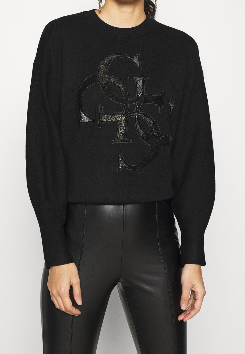 Femme portant un pull noir avec un logo en lettres brillantes superposées et un pantalon en cuir noir taille haute, debout devant un fond blanc.