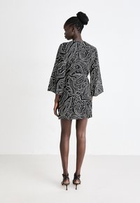 Abito nero corto con motivo paisley bianco a spirale, maniche a campana e una forma aderente, abbinato a tacchi neri con cinturini.