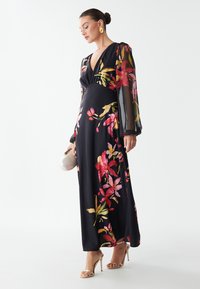 Abito lungo nero con stampa floreale colorata, scollatura a V profonda, maniche lunghe trasparenti e gonna fluida. Accessoriato con una pochette e tacchi a strappo.