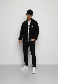 Jachetă neagră din bumbac cu un patch cu logo, tricou alb cu design grafic, blugi negri și adidași albi. Accesorizat cu o căciulă neagră.