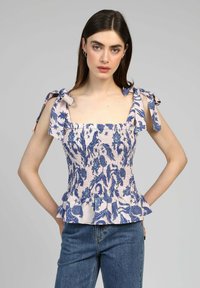 Haut peplum rose clair avec un motif floral bleu, décolleté carré et bretelles à nouer. Dispose d'un panneau smocké et d'un ourlet froncé. Porté avec un jean.