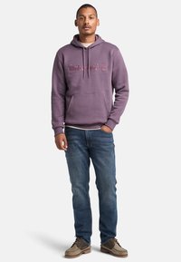 Felpa viola con logo "Timberland" ricamato, coulisse, tasca frontale; abbinata a jeans blu e calzature marroni.