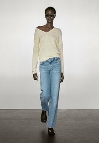 Maglione lavorato a maglia giallo chiaro con scollo off-the-shoulder e maniche ampie, abbinato a jeans a gamba larga blu chiaro e sandali neri.