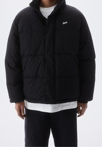 Doudoune noire avec fermeture éclair à l'avant, col montant et logo sur la poitrine. Présente une forme carrée et un rembourrage matelassé pour l'isolation.