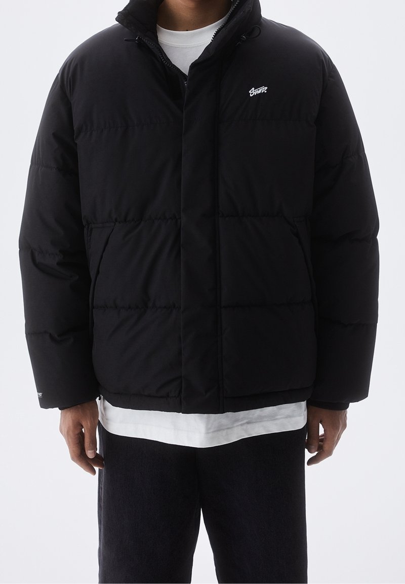 Doudoune noire avec fermeture éclair à l'avant, col montant et logo sur la poitrine. Présente une forme carrée et un rembourrage matelassé pour l'isolation.