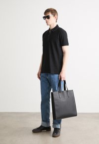 Polo nero con colletto zip, jeans blu e borsa a tracolla in pelle nera. Modello con occhiali da sole scuri e scarpe casual. Design minimalista.
