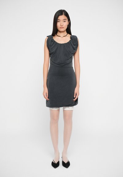 Robe grise ajustée avec décolleté drapé et manches courtes, portée avec un short à bord en dentelle. Le modèle porte des collants transparents et des talons noirs.