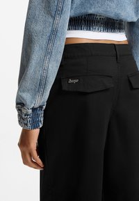 Τζιν τζάκετ σε ανοιχτό μπλε με σούρες στα μανίκια, συνδυασμένο με μαύρο παντελόνι cargo με δύο επίπεδες πίσω τσέπες και λογότυπο.