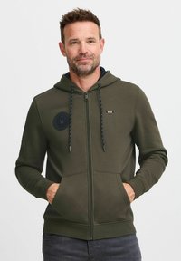 Sudadera con cremallera en verde oliva, fabricada en una mezcla de algodón, que presenta un diseño de parche redondo, capucha con cordones en azul marino y bolsillos frontales.