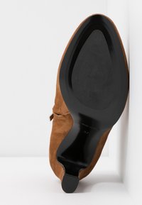 Brun ruskind højhælet sko med spids tå og sort sål, der viser en blød tekstur og minimalistiske designelementer.