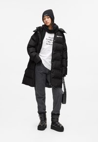 Vrouw met een zwarte beanie, sjaal, lange pufferjas, wit bedrukt T-shirt, grijze jeans en zwarte laarzen, die een zwarte handtas vasthoudt tegen een witte achtergrond.