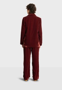 Aksamitny garnitur w głębokim burgundzie, składający się z długiego marynarki z klapkami i spodni z szerokimi nogawkami. Gładka faktura, dopasowany krój z miękkiego materiału.