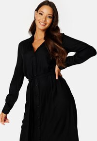 Abito camicia nero realizzato in tessuto morbido; maniche lunghe, chiusura a bottoni e vita con cintura per una silhouette aderente. Textura liscia e design sartoriale.