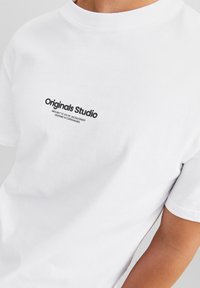 White cotton T-shirt featuring a crew neck and short sleeves. Black printed text reads "Originals Studio" on the chest.