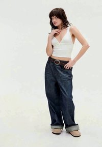 Fehér rövidített felső, sötétkék, bő szárú farmernadrág felgöngyölt szárakkal, barna bőr öv ezüst hardware-ral, és bézs papucsok. Egyszínű háttér.