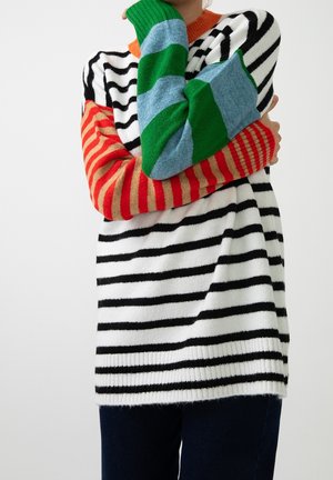 Personne portant un pull rayé noir et blanc avec une manche rayée rouge et orange, l'autre manche rayée verte et bleue, les bras croisés.