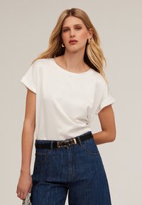 Top alb cu mânecă scurtă și decolteu rotund, purtat cu pantaloni din denim cu talie înaltă, care au o curea neagră cu accente metalice.