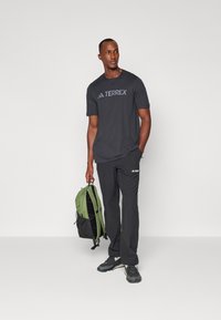 Sort T-shirt med "TERREX" logo, sorte bukser, grøn rygsæk og grå sportssko. Fokus på funktionelt outdoor tøj.