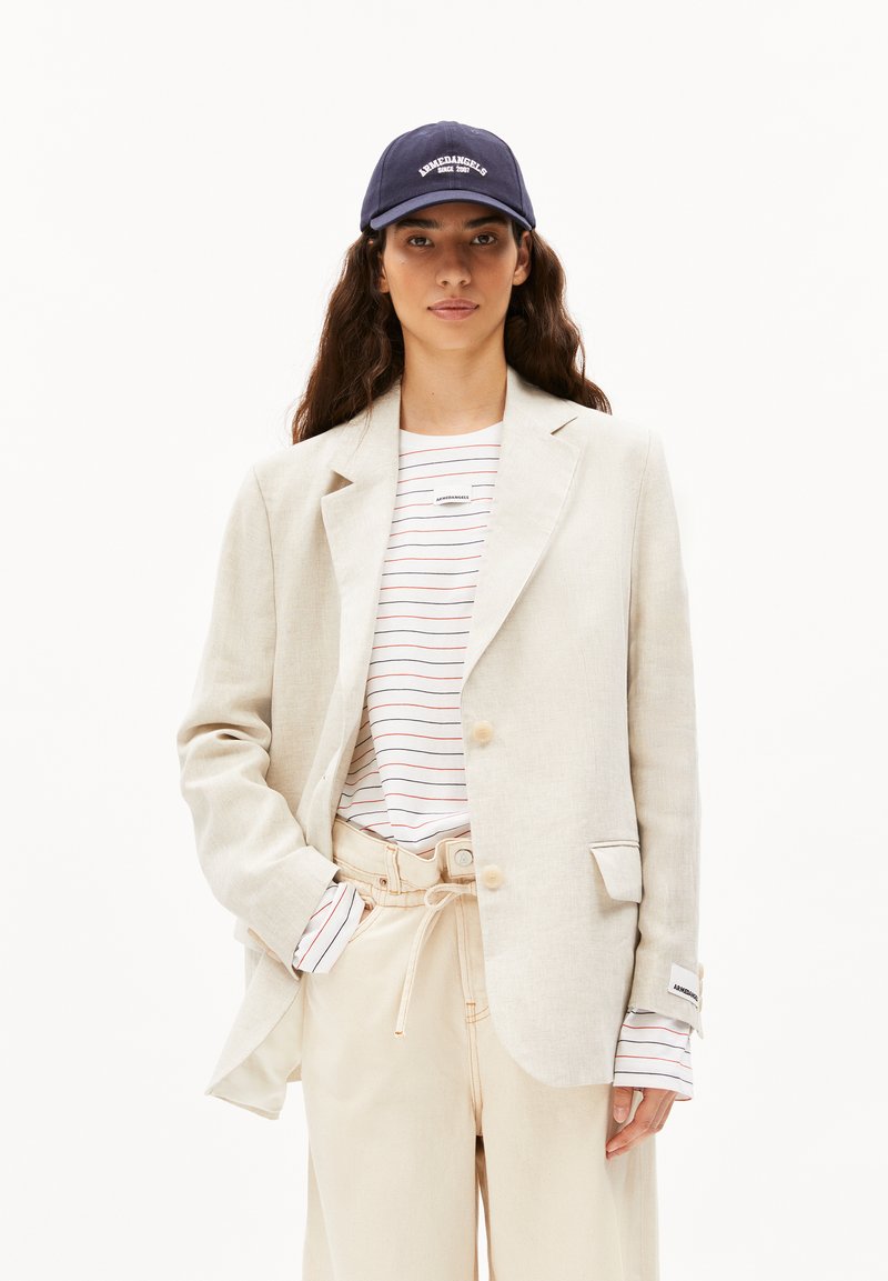 Blazer in lino beige chiaro sopra una camicia bianca a righe con linee rosse, abbinato a pantaloni crema e un berretto blu navy con testo bianco.