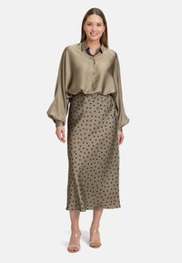 Blusa in satin oliva con una vestibilità ampia e chiusura a bottoni, abbinata a una gonna khaki a pois slim-fit. Sandali trasparenti con tacco.