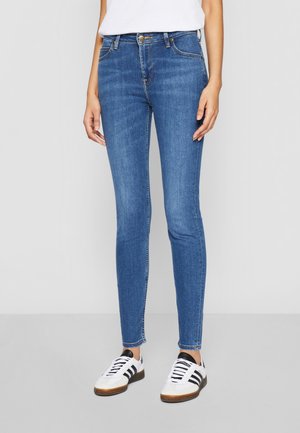 Jean slim bleu porté avec des baskets blanches à rayures noires et un haut blanc uni, debout devant un fond uni.
