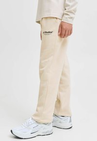 Beige sweatpants met een relaxte pasvorm, met zijzakken en het "JJ Studios" logo in het zwart. Gecombineerd met witte sneakers.