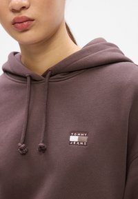 Hnědá bavlněná mikina s kapucí na stahovací šňůrku a malou vyšívanou nášivkou s nápisem "TOMMY JEANS" na hrudi.