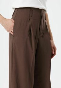 Pantalon marron taille haute à plis, avec poches latérales, en tissu lisse, présentant une coupe ajustée et structurée.