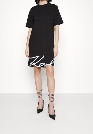 Femme portant une robe noire à manches courtes avec une inscription blanche à l'ourlet et des escarpins noirs à bouts pointus avec des lacets s'enroulant autour de la cheville, debout sur un fond blanc.