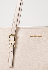 Beige leather handbag with a textured finish, featuring a gold-tone logo, adjustable strap, and buckle hardware.