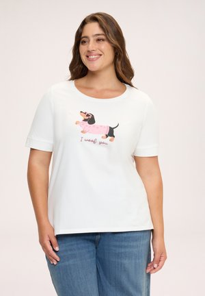 Lachende vrouw die een wit T-shirt draagt met een cartoon-teckel in een roze trui en tekst "I woof you,", gecombineerd met een blauwe spijkerbroek.