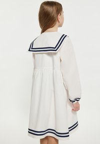 Weißes Kleid mit einem dunkelblauen Matrosenkragen und Ärmelaussetzern. Es hat eine geraffte Taille und einen gestreiften Saum und besteht aus einem weichen Stoff.