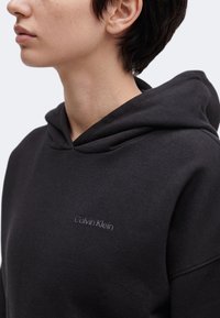 Άτομο που φοράει μαύρο φούτερ Calvin Klein, εμφανίζοντας το άνω μέρος του κορμού και το προφίλ του προσώπου μπροστά σε φόντο χωρίς σχέδια.