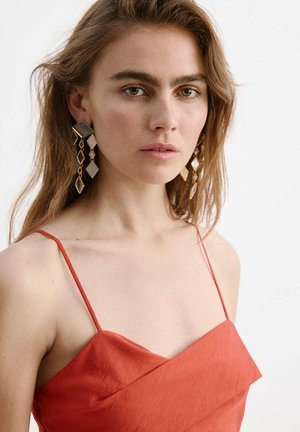 Jeune femme aux cheveux châtain clair porte de grandes boucles d'oreilles géométriques en or et une robe rouge sans manches sur un fond blanc uni.