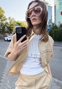 Camisa beige de manga corta con acentos de perlas, superpuesta sobre una camiseta blanca de canalé con un patrón de cuentas vertical; sosteniendo un teléfono inteligente negro.