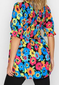 Kaleidoskooppinen kukkaolkaimilla varustettu paita, jossa on smokkikohouma, puffihihat, röyhelöhihojen päät ja sekoitus sinisiä, vaaleanpunaisia, oransseja ja keltaisia kukkia mustalla taustalla.