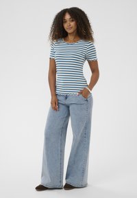 Gestreiftes Kurzarmoberteil mit wellenförmigem Ausschnitt, das blaue und weiße horizontale Streifen zeigt; kombiniert mit hellblauen Jeans mit weitem Bein.