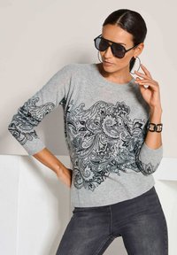 Frau trägt einen grauen Pullover mit schwarzem Paisleymuster, schwarze Jeans, große schwarze Sonnenbrille, Creolen und ein schwarzes Armband.