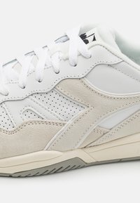 Sneaker bianco con una combinazione di pelle liscia e suede beige. Il design presenta accenti perforati e lacci piatti bianchi.