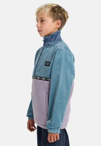 Pullover in corduroy azzurro chiaro e pile viola con colletto alto, bande nere con motivo e tasca frontale. Maniche lunghe con polsini.