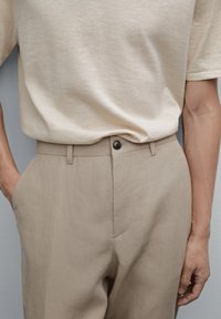 Beige Kurzarmhemd, kombiniert mit hellbraunen Hosen, die über einen Knopfverschluss und seitliche Taschen verfügen. Der Stoff wirkt weich und strukturiert.