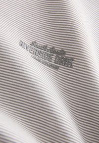 Stribet stof i lysegrå og hvid med en glat tekstur. Præget med mærkenavn og produktbeskrivelse i mørkegrå.